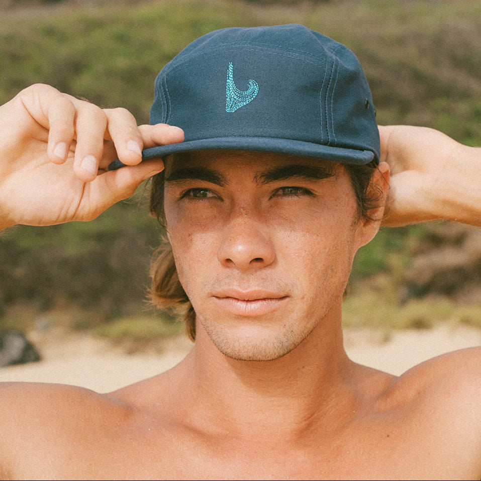A person wearing a blue five-panel hat with a teal embroidered logo on the front, adjusting the hat with one hand while looking to the side.