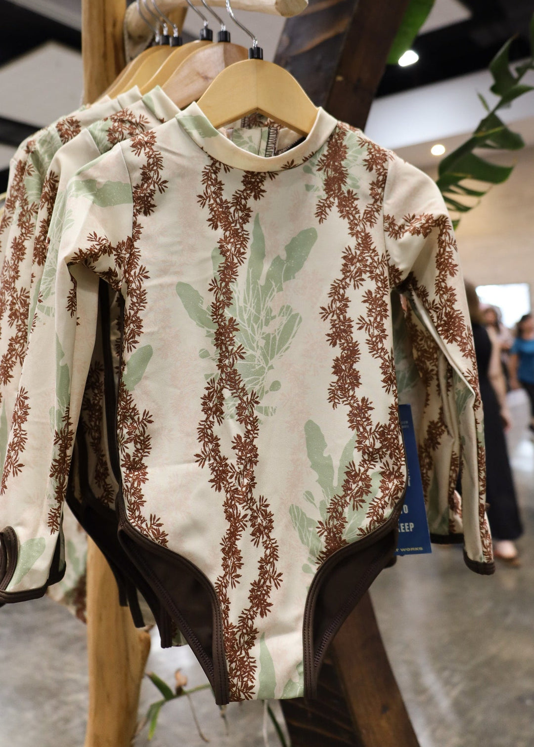 A long-sleeve baby bodysuit with a botanical limu seaweed pattern in brown and sage green on an off-white background, hanging on a wooden hanger on a clothing rack inside a store.