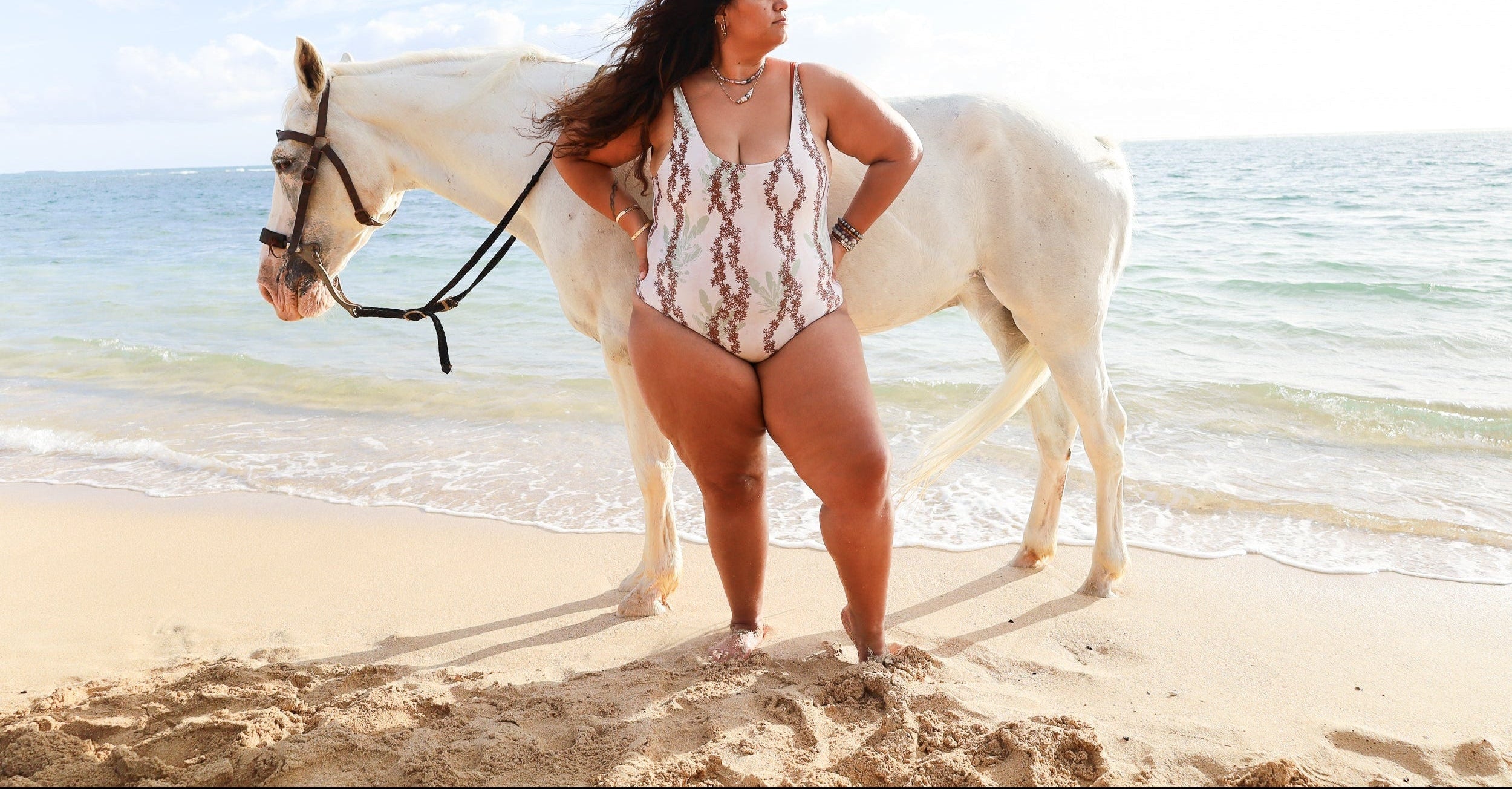 A woman stands on a sandy beach next to an ocean, wearing a white one-piece swimsuit with a brown pattern. A white horse is standing beside her, and the sea and sky are visible in the background.