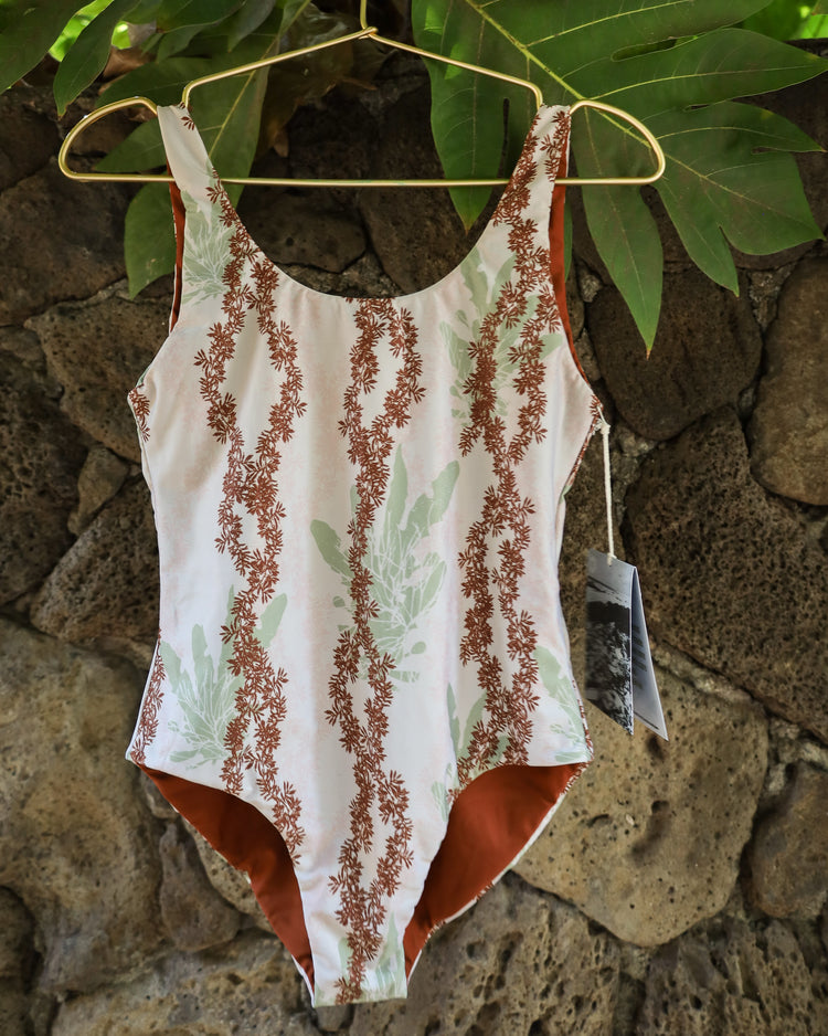 A one-piece swimsuit with a cream white base, brown floral lei limu seaweed patterns, and light green leaf motifs, hanging on a gold hanger against a rustic stone wall with green ulu leaves.