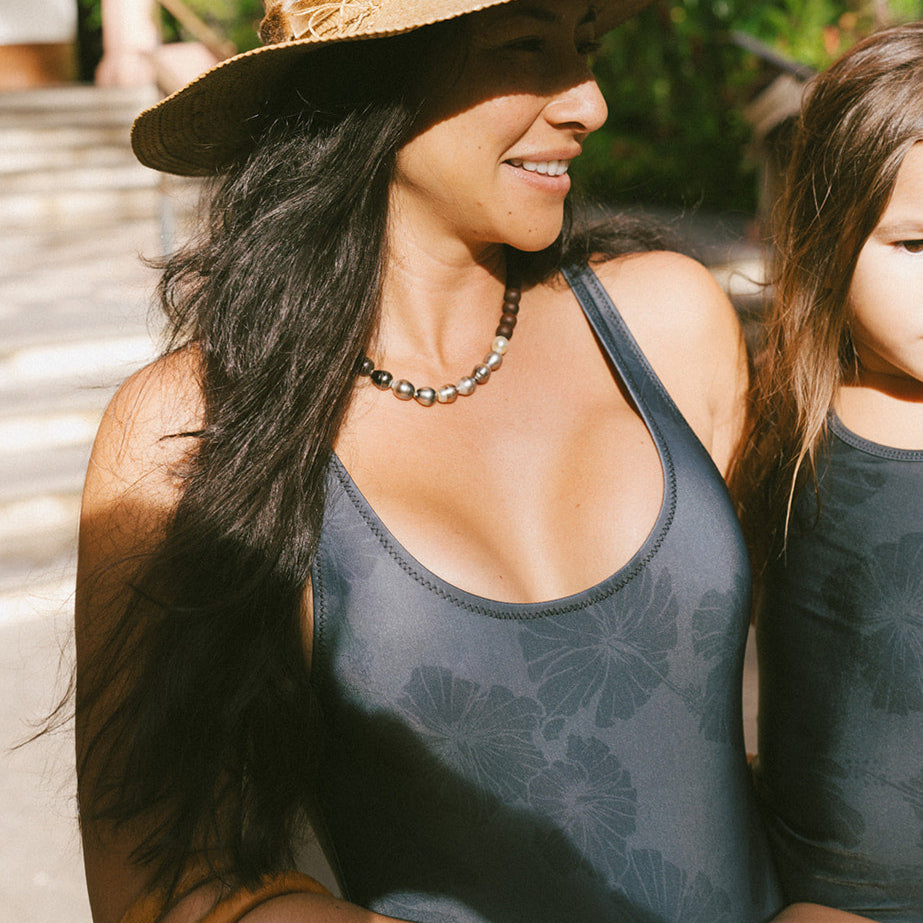 woman wearing a black on black italian made swimsuit with subtle hibiscus flowers carrying her daughter in the matching suit. its total luxury.